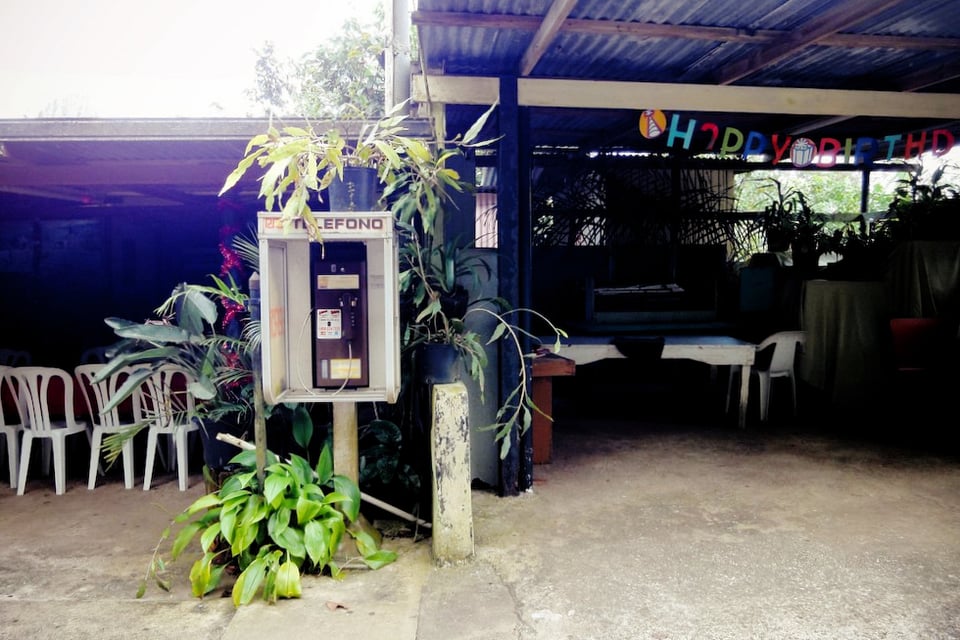 An open air structure with an old pay phone labeled "Telefono" in front, with tropical greenery growing around it. In the background, white plastic chairs and a "Happy Birthday" banner suggest a festive gathering.