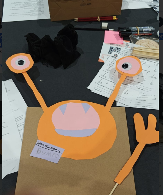 A puppet made of paper sits on a black tablecloth at a convention. The puppet has googly eyes and giant eyestalks, and is wearing a sticker that says "Hello, my name is BUMPO."