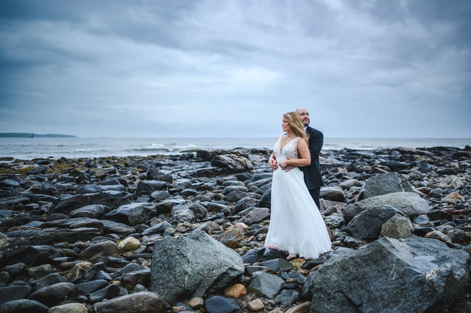 Bride and groom embrace each other and gaze off into the distance on a rocky new england beach during a coastal storm