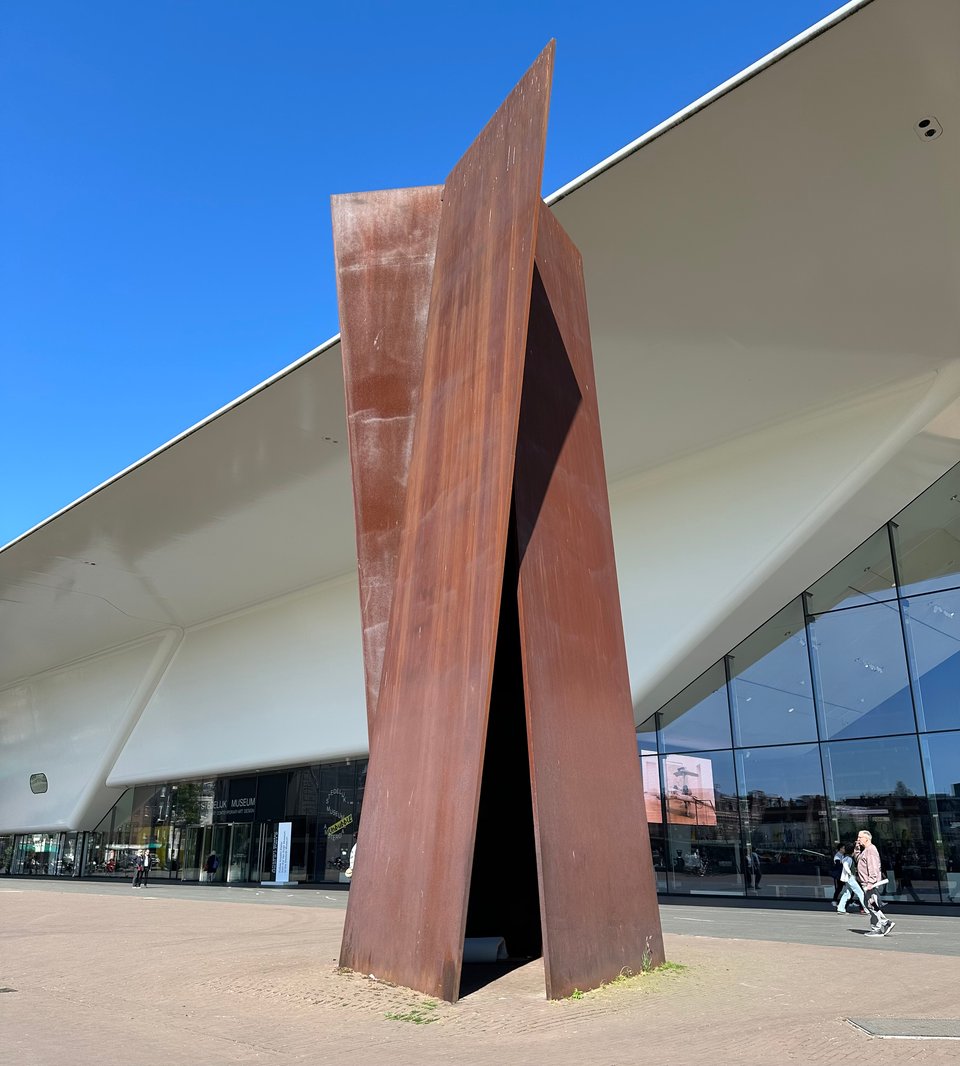 photo of Sight Point, a sculpture by Richard Serra, consisting of 3 17-ton slabs of stainless steel balanced against each other to form a pyramid. There's a thin foam mattress folded up inside at the base, where some poor soul must sleep for shelter