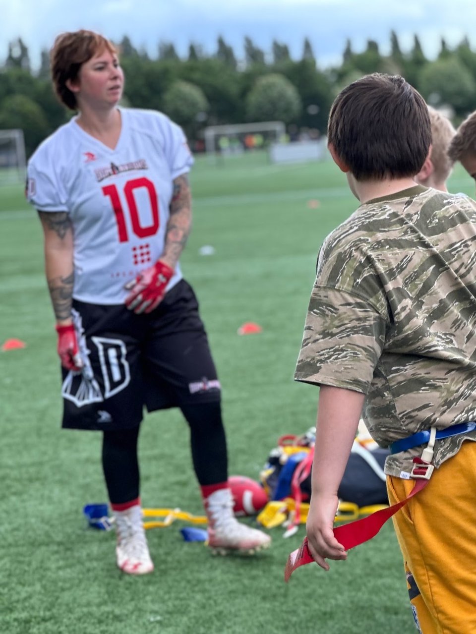 A woman explains the rules of American Flag Football, children seen from behind look and listen.
