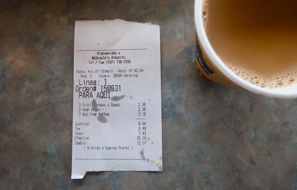 A fast-food receipt with coffee stains sits on a table next to a cup of light brown coffee. The receipt lists breakfast items with prices in Spanish.