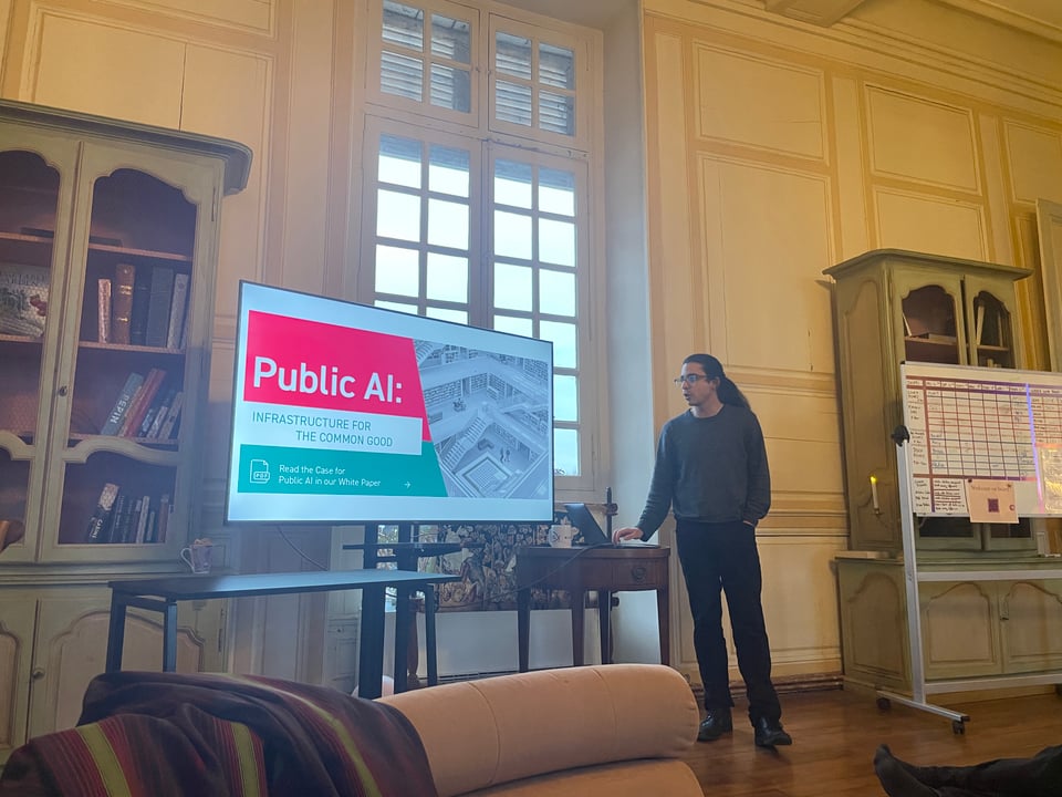A man with long hair and glasses gives a presentation entitled "Public AI Infrastructure for the common good"