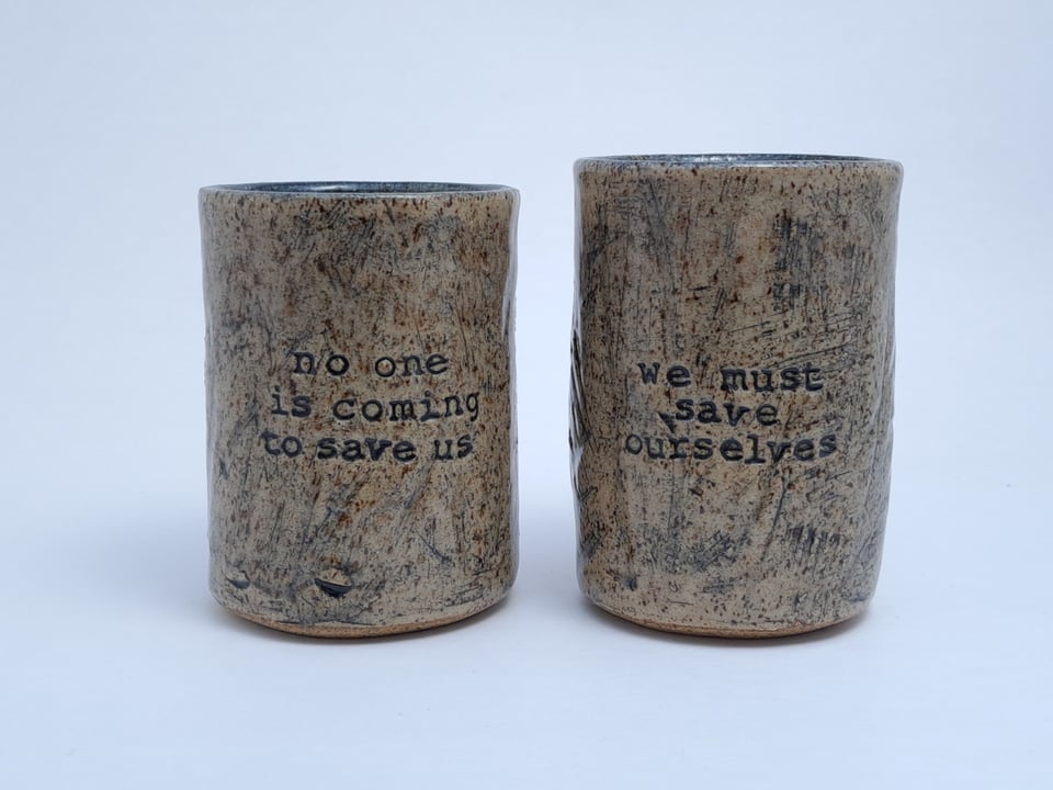 Two ceramic cups stamped with "no one is coming to save us" and "we must save ourselves"
