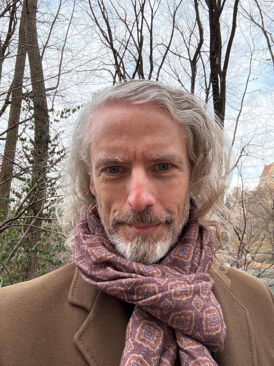 photo of bearded white man in a brown overcoat and purple-ish patterned scarf looped around his neck. He has long hair and is smiling at the camera. Central Park trees are in the background