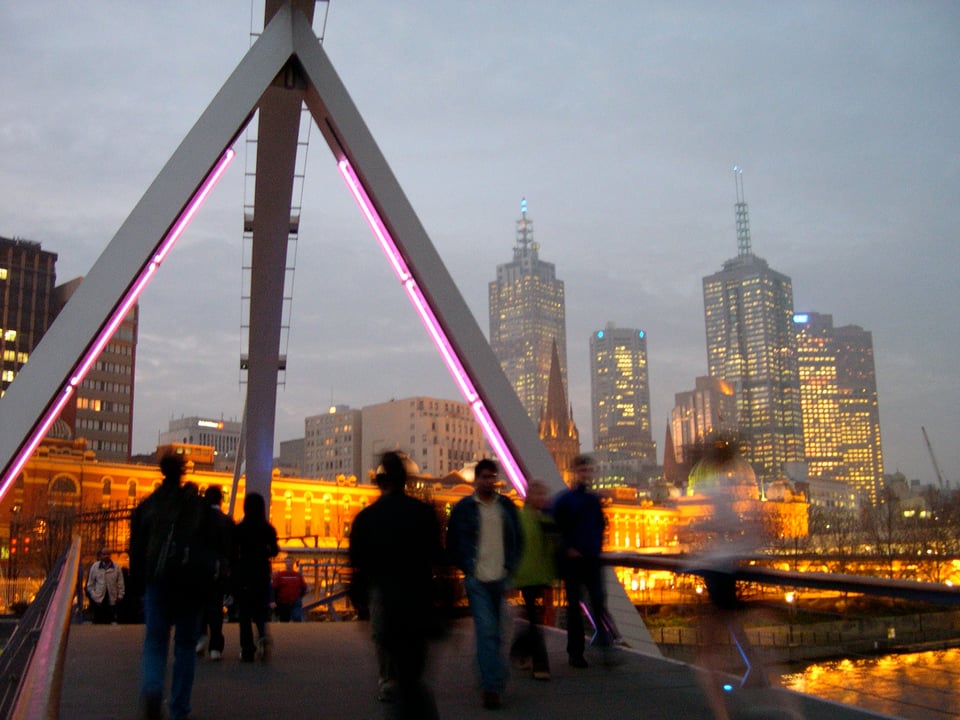 A murky grey sky at dusk. Under it Melbourne's city centre with warm, golden lights. In the foreground a bridge with people but the people are like shadows and silhouettes. One of them, on the right hand side, is like a blur, like a ghost.