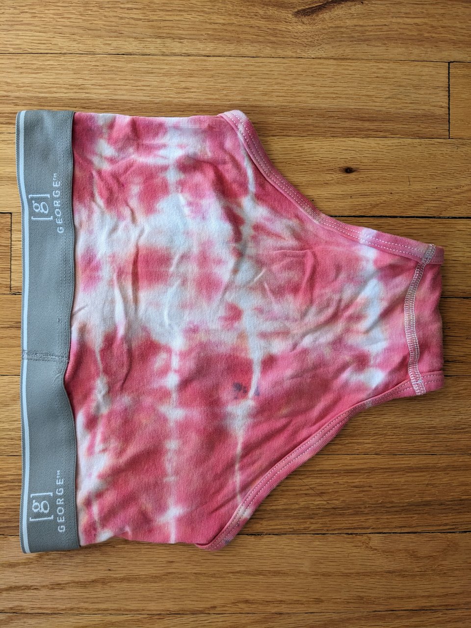 A pair of tie-dyed briefs, reddish striped with a gray waistband, against a hardwood floor. The briefs are showing backside up and are tilted to a "side" view with the waistband facing the left of the pic.