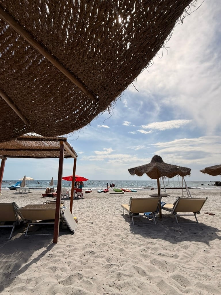 A sandy beach viewed from beneath woven sunshades, with loungers and parasols facing the sea and kayaks lined up near the water.