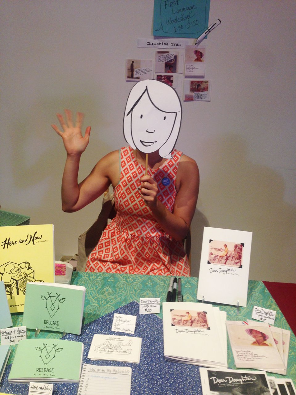 Christina (holding up a comic Stina mask) waving hello from behind a zine fest table
