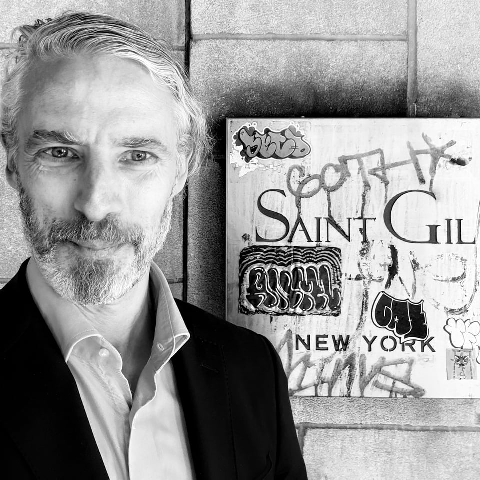 B/W portrait photo of white man in suit & dress shirt (no tie), standing next to a metal sign that reads SAINT GIL; it is covered in graffiti & stickers