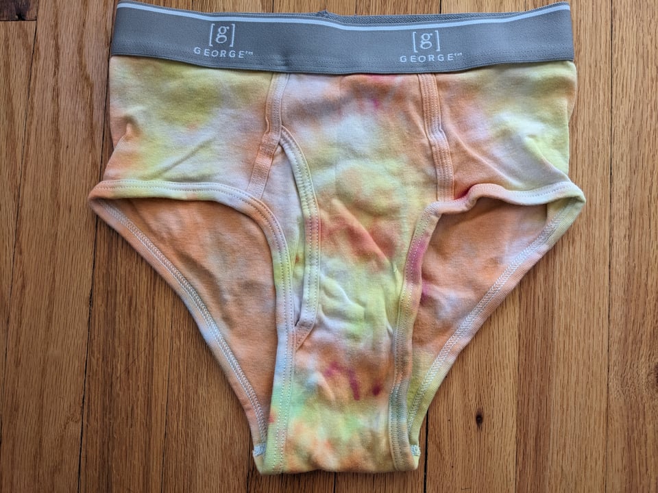 A pair of tie-dyed briefs, mottled yellow and orange with a gray waistband, against a hardwood floor.