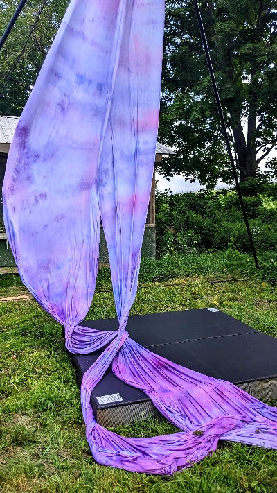 A long double length of silky fabric hung from a rig and billowing in the breeze; it is a watercolor tie dye ombre effect of purple to blue to pink above