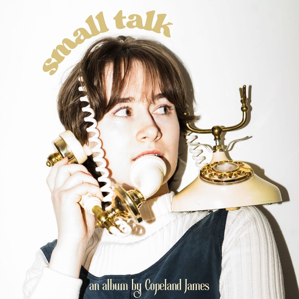 Album cover for "Small Talk." Copeland James, a white person with short brown, is looking up and to the left with a vintage telephone perched on their left shoulder. The phone cord is wrapped around their head as they hold the hold up to their right ear. The words "Small Talk" curve around the top of their head in a gold, bubble-letter print. At the bottom of the image are the words "an album by Copeland James."