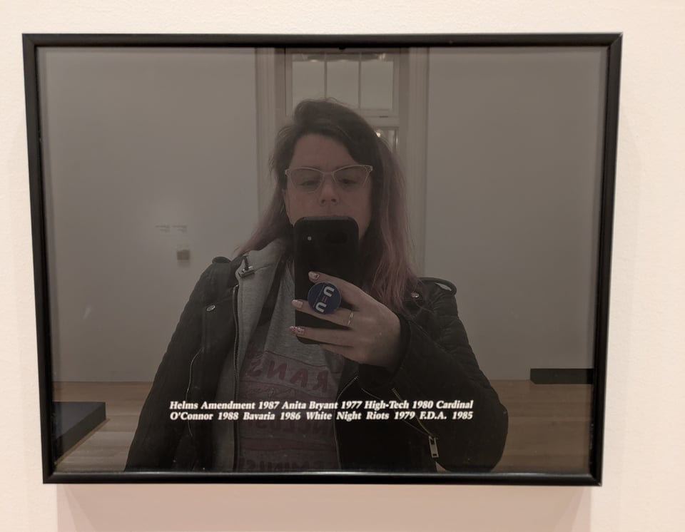 A black framed image with words and dates across the bottom, that is so reflective the photographer can be seen clearly in its mirror-like surface.
