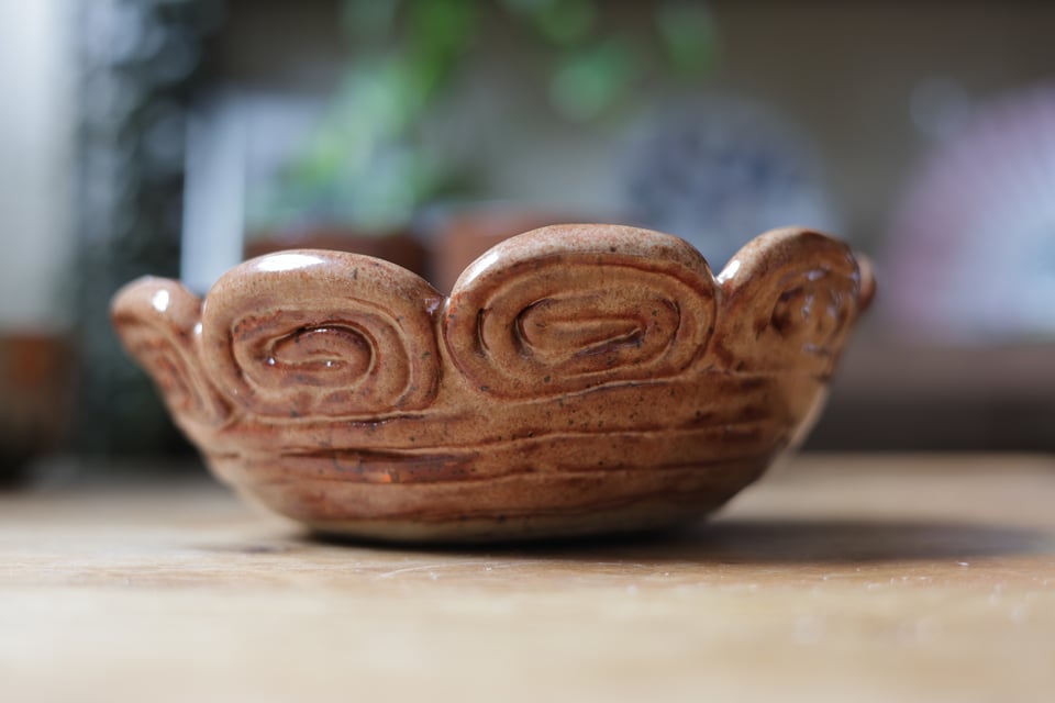 A brown ceramic bowl made of clay coils, some horizontal to build the base with decorative spirals on top.