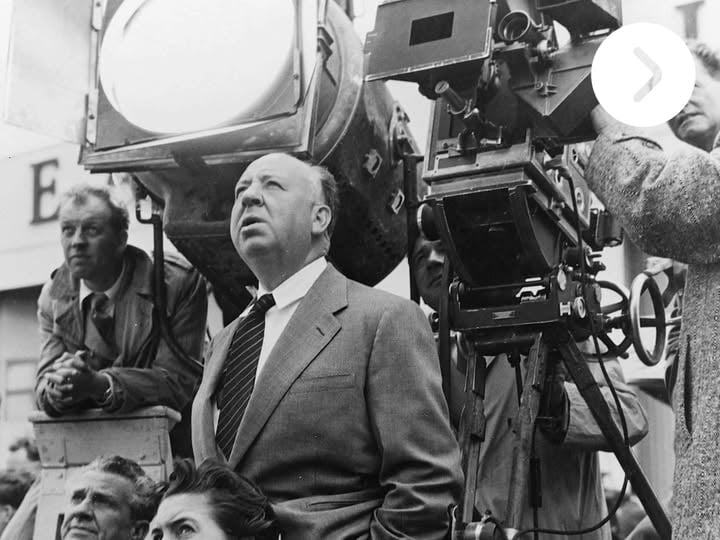 Alfred Hitchcock directing on set, surrounded by crew members and cameras, looking off in the distance