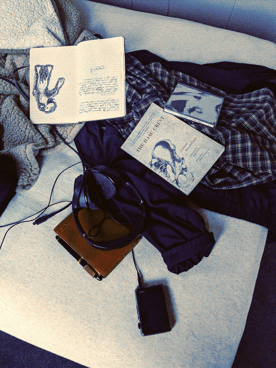 A patterned shirt, a jacket, a pocket notebook, an mp3 player, a pair of headphones, a paperback novel, and an open moleskin all strewn across an unmade twin-size bed.
