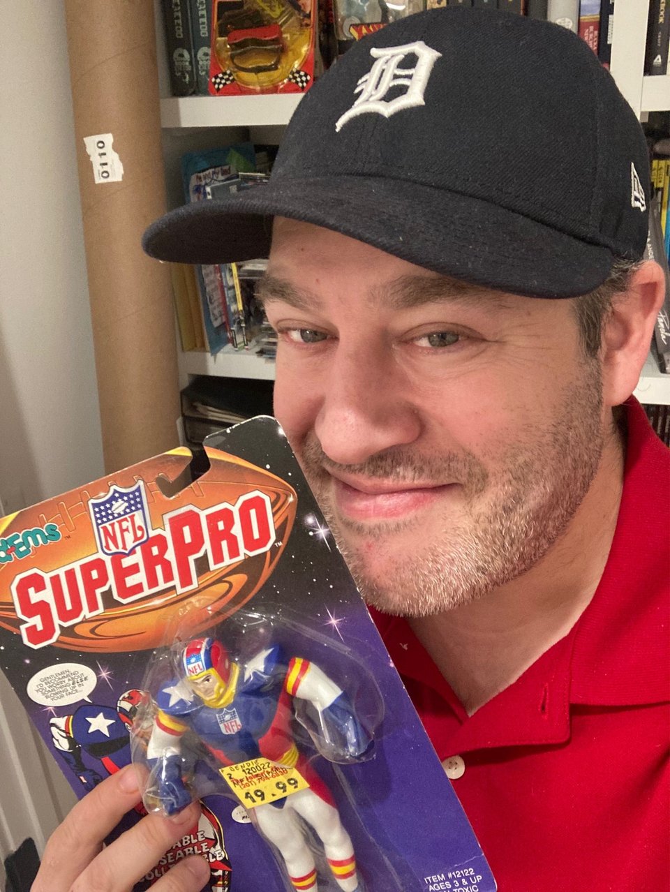neil in a red polo shirt and a navy detroit tigers ball cap, holding a NFL superpro bendy toy in its package