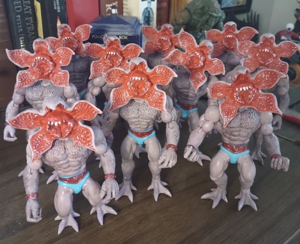 A group of pale gray creatures with flower-like heads stands on a wooden desk. There's ten Demogorgons on the desk, all with their distinctive opening-petals head that reveals red flesh and lots and lots of teeth. But they also all have the equally distinctive He-Man/ Masters of the Universe physique, with broad, muscley chests and swollen biceps and thighs.