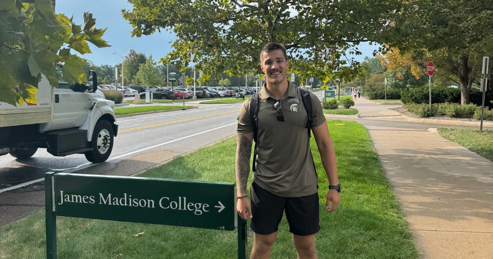 A young man stands smiling beside a green sign that reads 'James Madison College' on a grassy area next to a road. A white truck is visible driving past in the background under leafy trees.