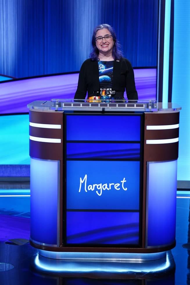 Your author, an anglo woman in her 40s with dark purple hair and wearing a space-themed dressed, grins like a maniac behind a Jeopardy! podium with "Margaret" written on the screen. Although it is not visible, the production team put me on a very high lift so that more of the dress would show. My blue/purple/black theme matches the Jeopardy! set very well, if I do say so myself.