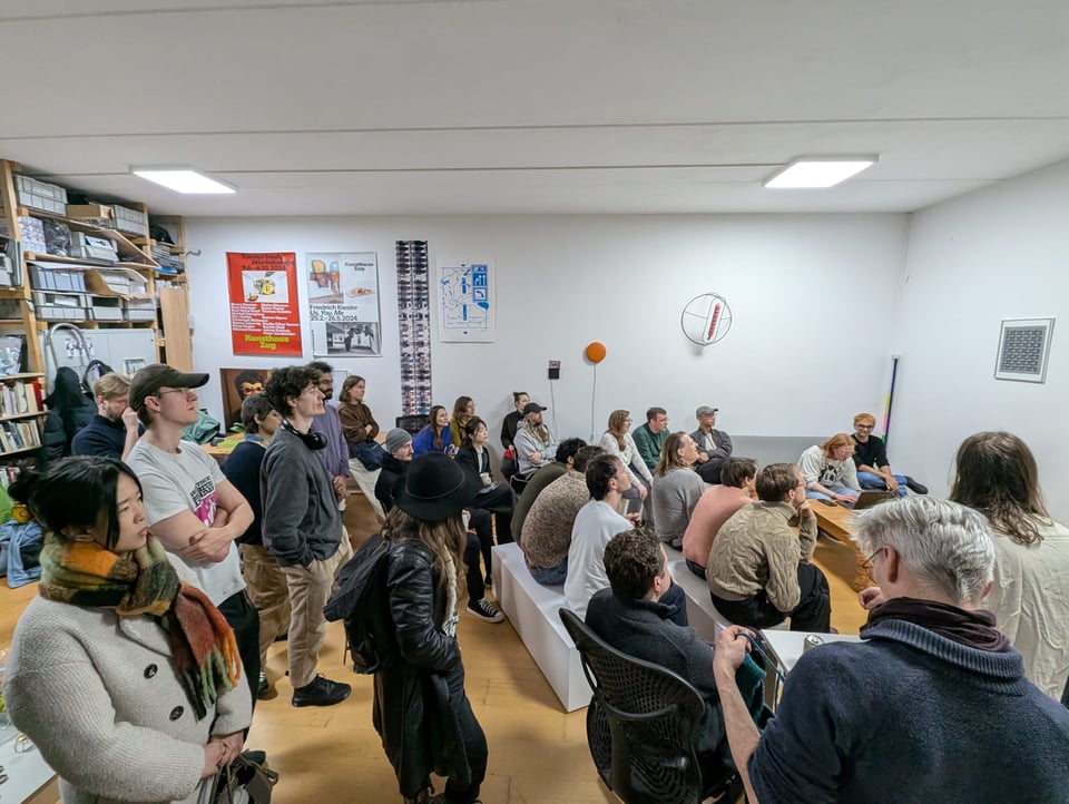 A group of people cluster around an off-frame TV in an expanding ring of couches and benches. Some at the back are standing, and listening to a seated speaker in a white t-shirt.