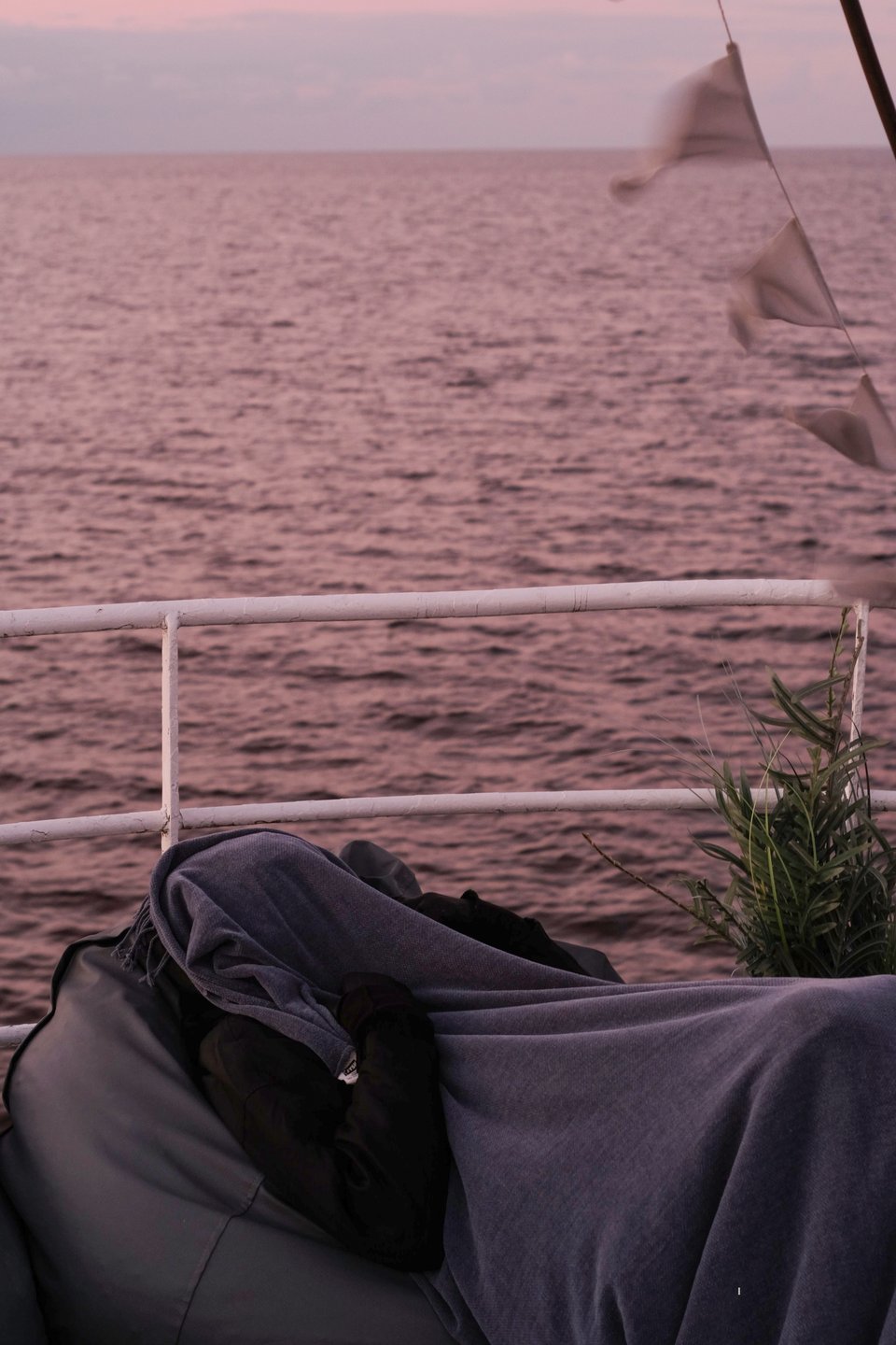 A photo of a person on a boat at sea, sitting in a bean bag, with a coat and gloves, protecting their face with a blanket. To the side, a flag garland can be seen fluttering in the wind.