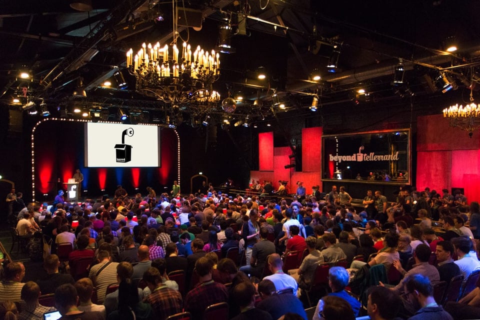 A photo showing the sold-out theatre of beyond tellerrand in Düsseldorf with 500 attendees in front of the stage. You can see the stage and a chandelier on the ceiling. The room is lit in mostly blue and red colours. A big mirror on the right side says beyond tellerrand.
