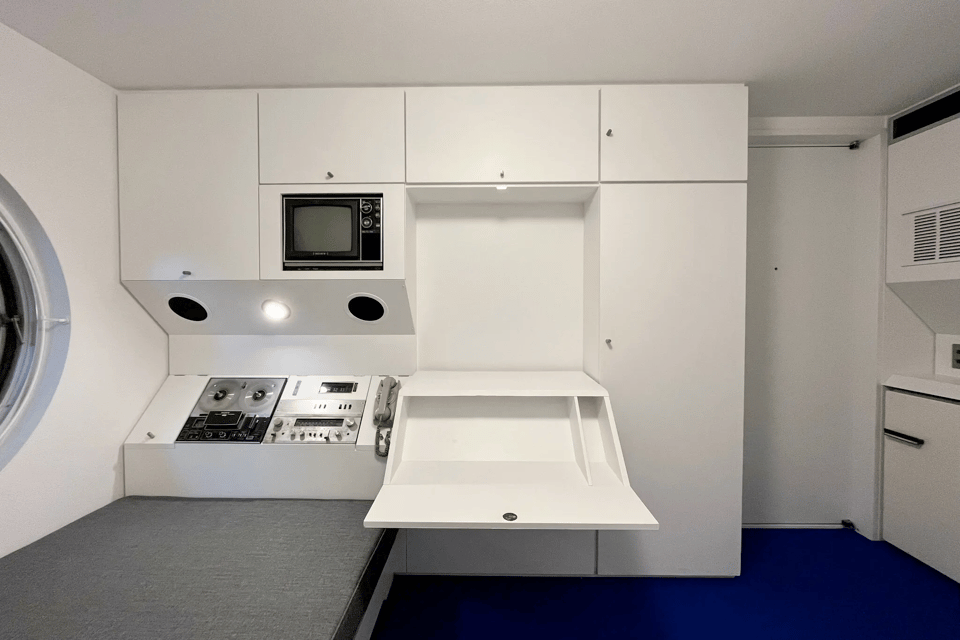 A photo of the inside of one of the capsules. You see, on the far left side, part of the round window, and beneath the windows a gray cot for sleeping. At the head of the cot is an angled white wall unit that has embedded in it a reel-to-reel tape player. Above that are a light and two speakers, and above those, a television set. The white wall-unit continues to the right, where there is a fold out desk that is folded out. On the far right side is a wall with more white units. The floor is black. It has a look of something you'd see in a science-fiction movie, almost as if it were the inside of a spaceship.