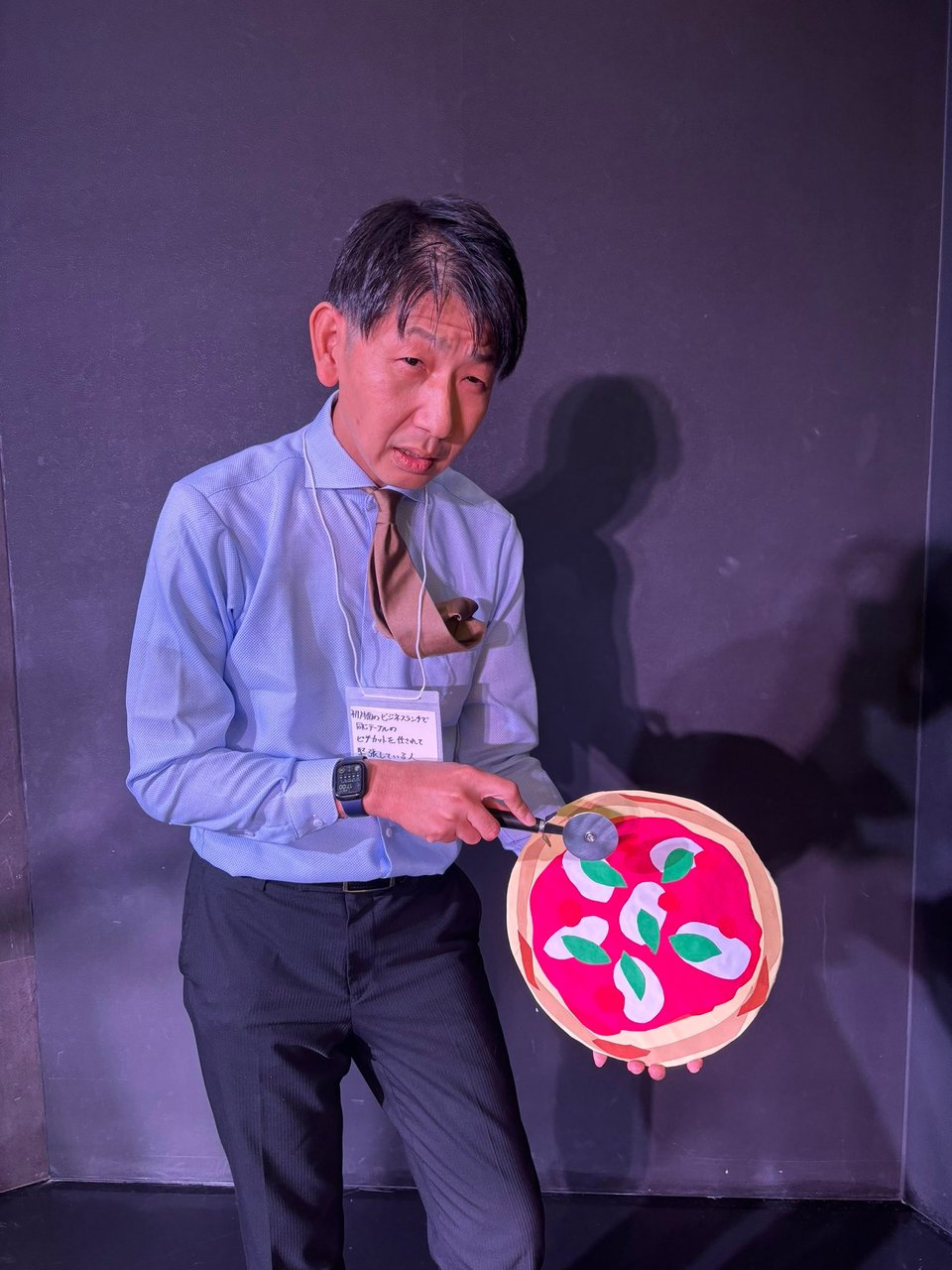 A man dressed as a salaryman holds up a fake pizza and pizza cutter