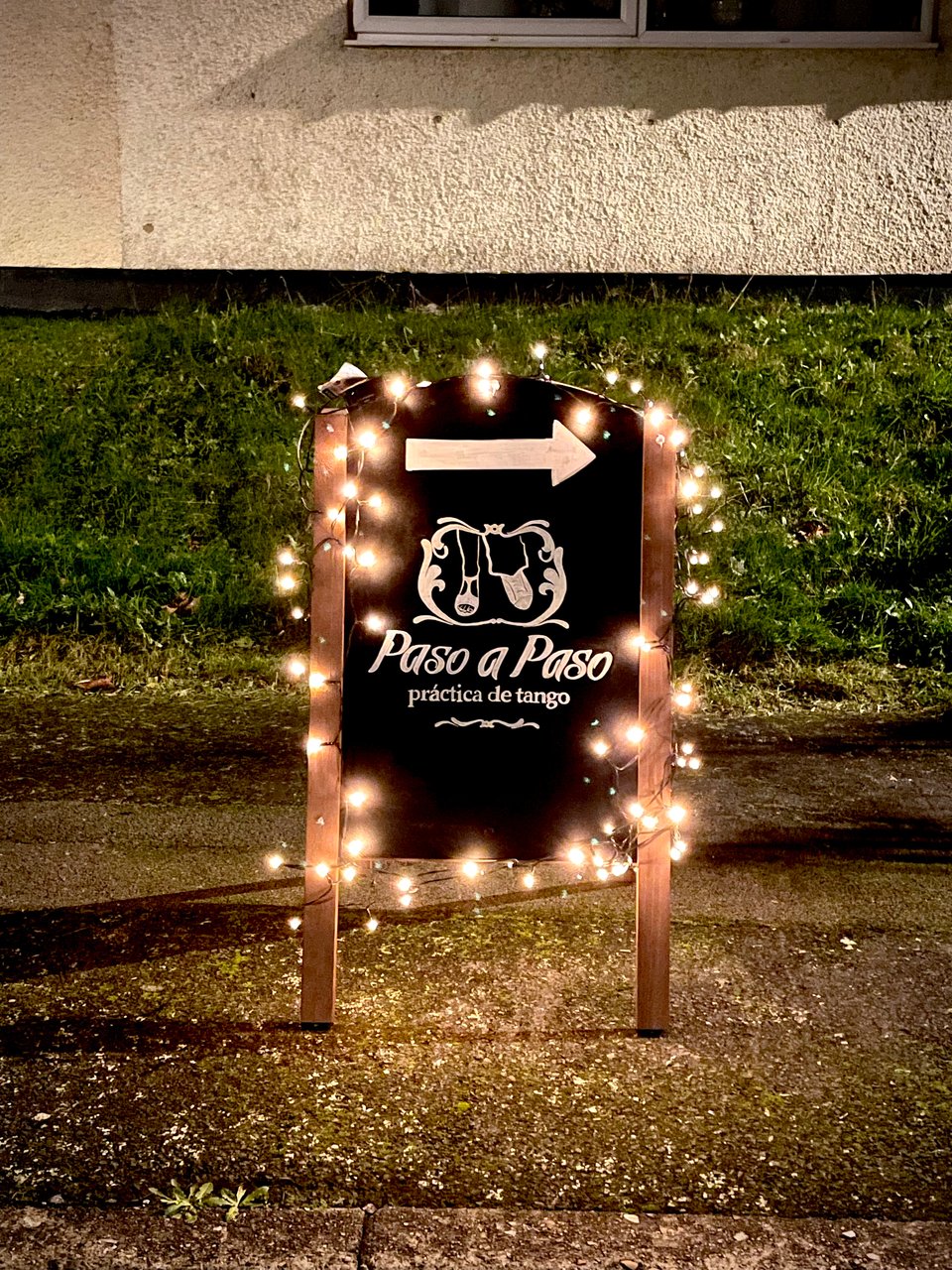 Pavement blackboard sign with fairy lights around its frame. Painted in white at the centre, the lodo of "Paso a Paso Practica de Tango" with an arrow on top point to the right"
