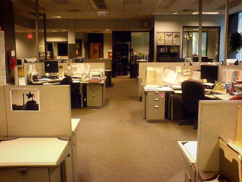 Empty newsroom under yellow fluorescent lights. Papers and personal belongings are scattered over desks; one hopes everyone will be back tomorrow.