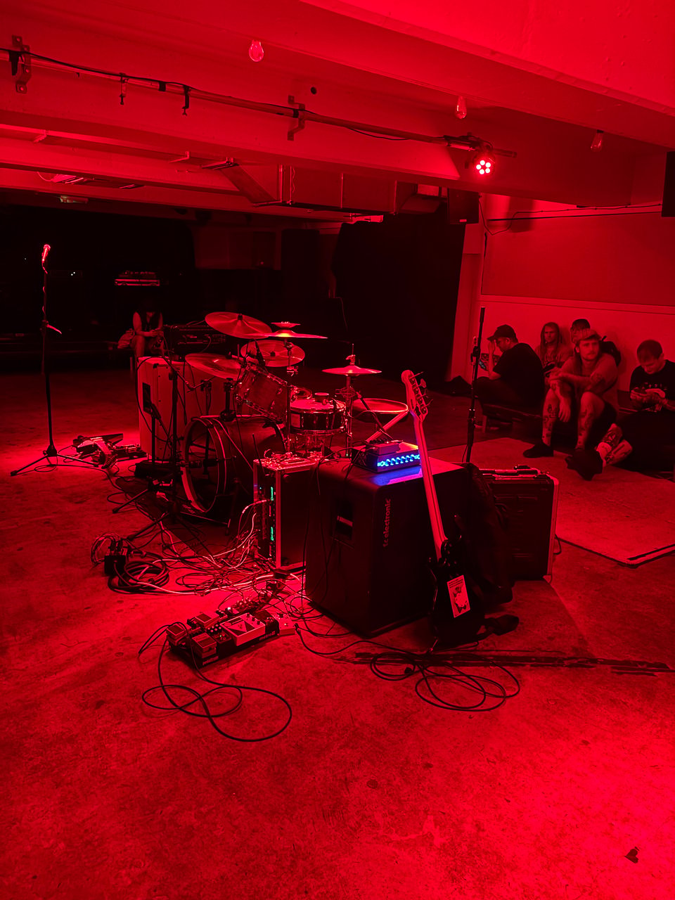 A dimly lit music venue illuminated in red, featuring a drum set, guitar, and various equipment, with a small audience seated in the background.