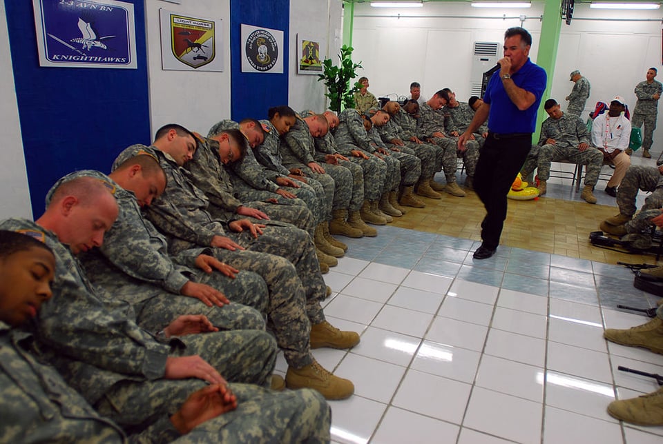 A roomful of uniformed soldiers with their eyes closed leaning on the shoulder of the person beside them, while a man speaks into a microphone.