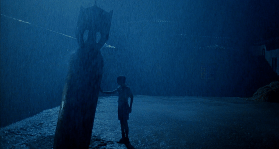 A young boy touches an unexploded missile standing upright in a bare, rain-soaked courtyard. The light is very blue. This is a still from The Devil's Backbone.