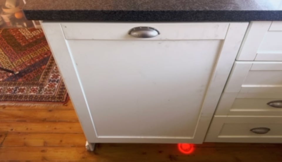 A built in dishwasher is closed and running with a red light on the floor to show that it is on.