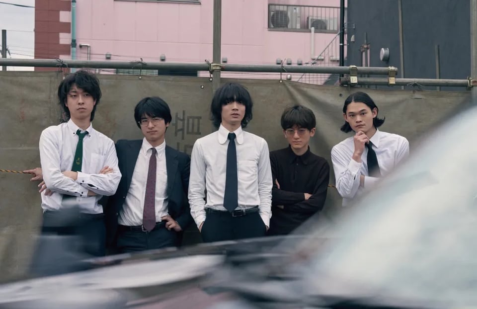 Five young men in white dress shirts and dark ties stand shoulder to shoulder against a weathered construction tarp, posing with crossed arms and serious expressions, while a pink building and overhead utility poles rise behind them and a blurred vehicle cuts across the bottom of the frame.