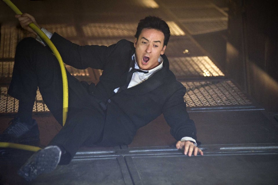 John Cusack wears a natty black suit and lays on the ground where he apparently just fell down. His mouth is wide open.