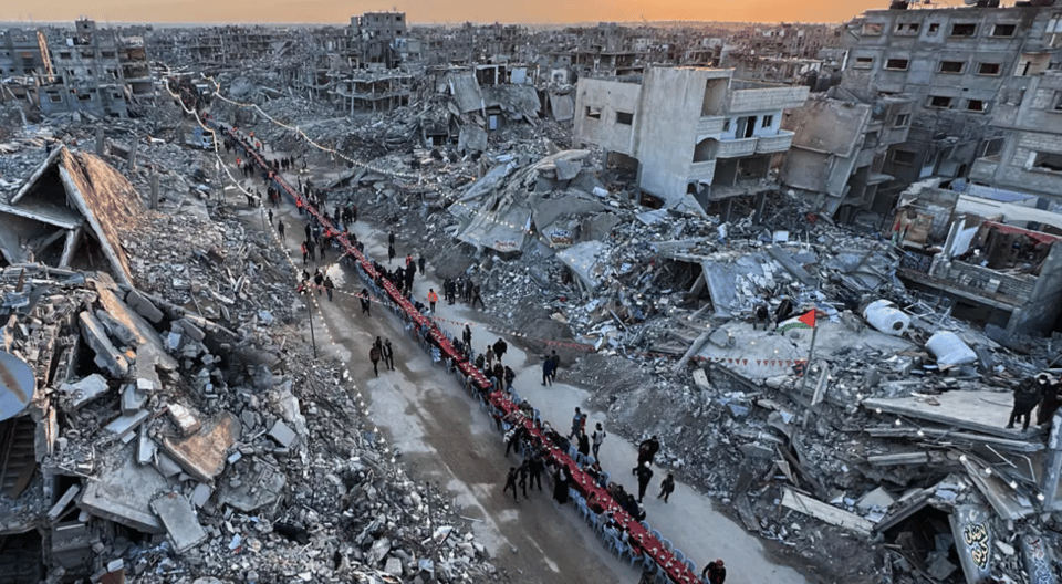 Amidst vast destruction of demolished concrete buildings, a very long table is set down the middle of the empty space that was presumably once a road