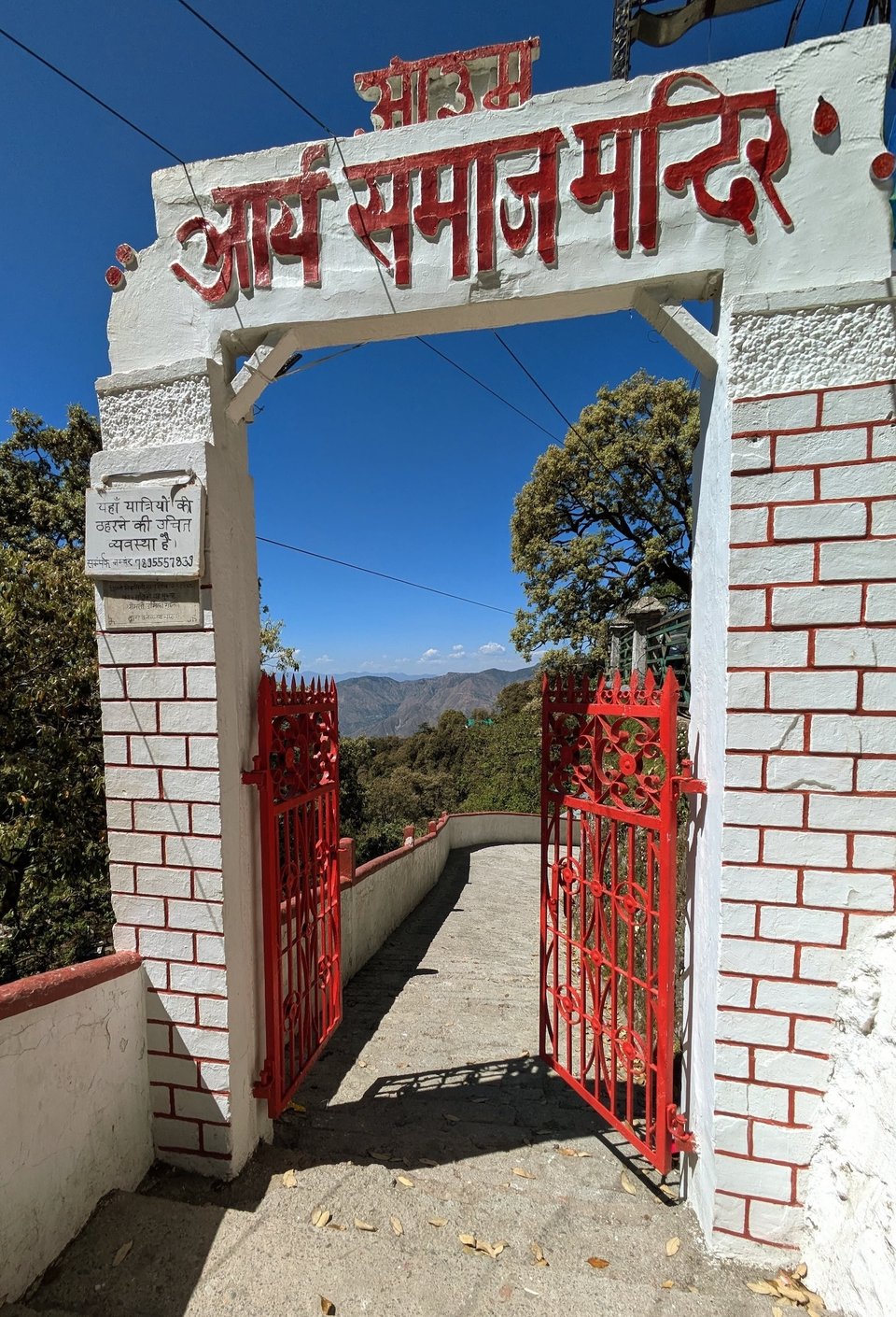 An image of a bright red metal gate made of sculpted rods beneath a sign that reads Arya Samaj Mandir (Arya community temple). The walls holding the gate are exposed brick walls than are painted off-white with red accents matching the gate. The pathway curves towards the end and there are mountains in the background. These are mountains at the foot of the himalyan range