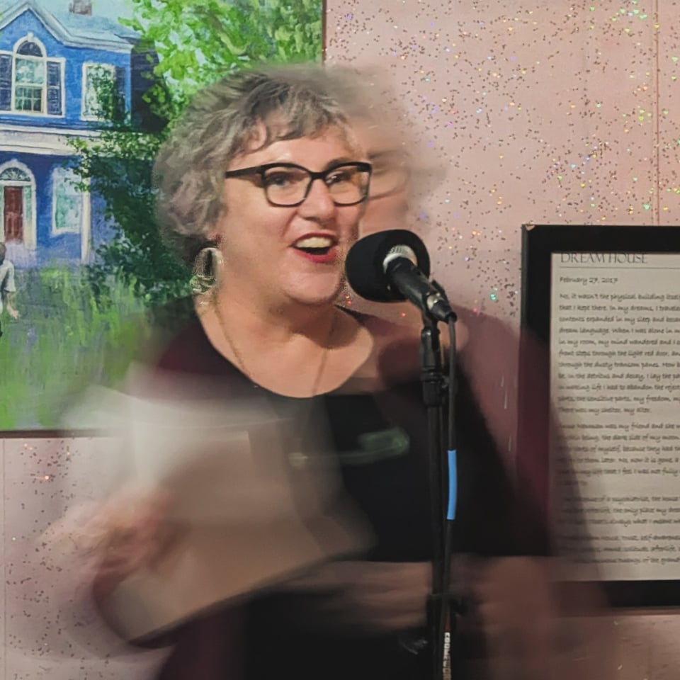 A white woman with glasses and dangling earrings is reading at a microphone. Her mouth is open and she's smiling as she reads. Due to the type of shot, she and her papers are mostly blurry, except for her face.