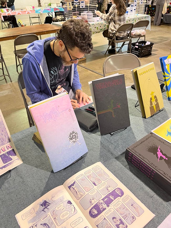 An artist sitting behind a table, drawing. There are comics on the table.