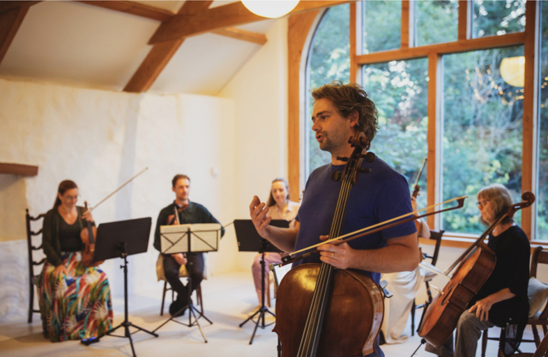 String quintet playing in a room lit by a large window overlooking a garden.