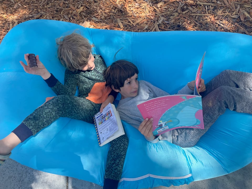An image of two school-age boys, brothers, sitting outside on an inflatable blue beanbag chair, reading books in the afternoon sun.