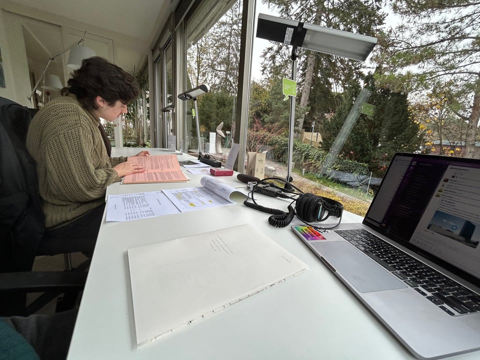 A person in a brown sweater, sitting at a desk looking at papers. In the foreground there is a computer screen open. Out the big windows are trees.