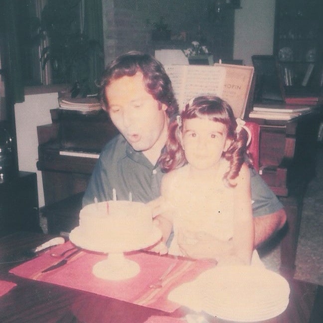 Man blowing out birthday candles while holding young daughter in pigtails, in front of piano, with open Chopin sheet music in background, 1970s