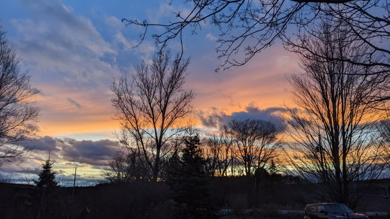 a vivid peach-gold sunset streaked across equally-vivid winter-blue sky, smeared and dappled with clouds, framed by dark bare trees and hills in silhouette