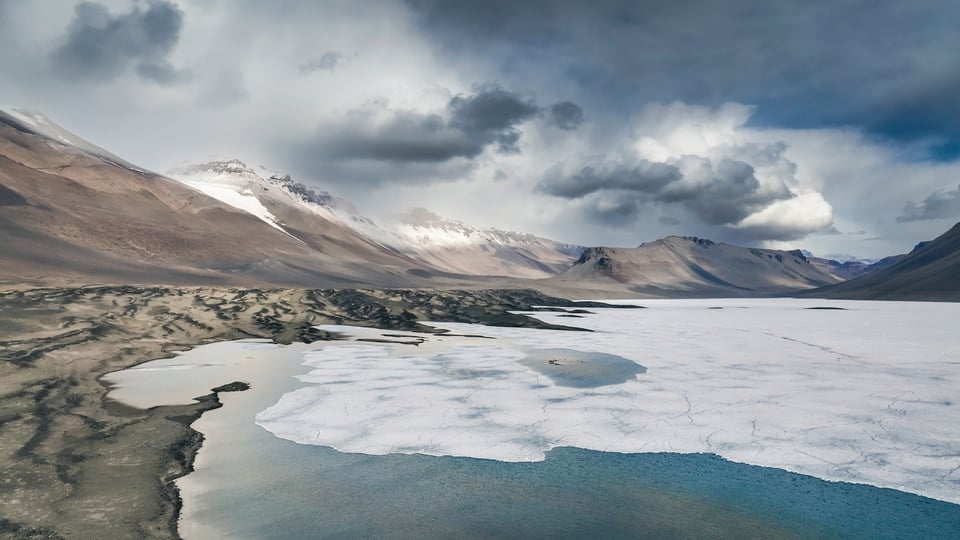 A photo of the McMurdo Dry Valleys in Antarctica, showcasing a partially-frozen lake, a wide range of multi-colored mountains, and stormy clouds.