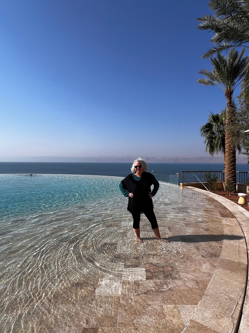 Woman with gray hair, black jumper, and capri leggings with her hands on her hips, standing in a beautiful round, shallow pool, with a body of water behind and land in the distance, under a bright blue sky, with palm trees on the right side of the pool