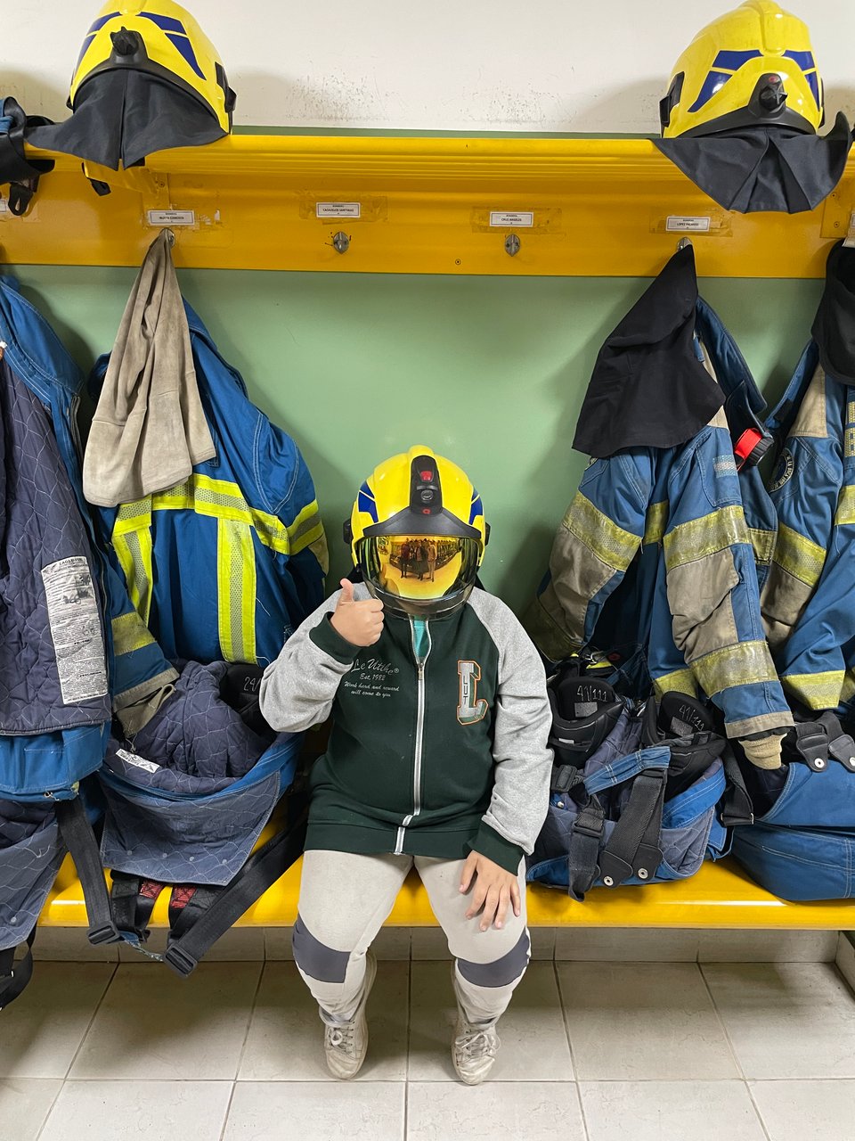 A young boy is seated on a bench wearing a reflective fire helmet. He is surrounded by fire fighting uniforms hanging on pegs.